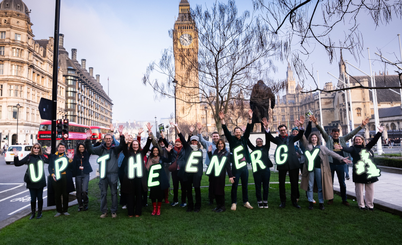 A group of around 20 people standing together in Parliament Square, London, with Big Ben in the background. They are holding up glowing green letters that spell out 'UP THE ENERGY' and raising their hands in celebration. The photo is taken at dusk with a red London bus visible in the background.